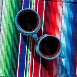 May include: Two blue ceramic mugs with white polka dots and handles, filled with a dark liquid, sit on a colorful striped blanket. The blanket features red, green, blue, and white stripes, creating a vibrant background.