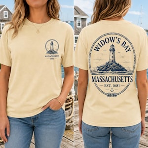 Könnte beinhalten: Ein cremefarbenes Kurzarm-T-Shirt mit einer kleinen Leuchtturm-Grafik auf der linken Brust und dem Text Widow's Bay Massachusetts 1681. Die Rückseite zeigt eine große, von einem Seil umrahmte Leuchtturm-Illustration mit demselben Text.