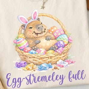 Könnte beinhalten: Cremefarbenes T-Shirt mit einem Capybara mit Hasenohren in einem Korb voller Ostereier. Der Schriftzug "Egg-stremely full" ist in lila gehalten. Eine silberne Halskette mit Sternanhänger liegt auf dem Shirt.