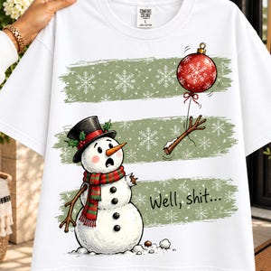 May include: A white short-sleeve t-shirt featuring a graphic of a surprised snowman wearing a black top hat and a red and green striped scarf. The snowman is missing an arm, which is attached to a red Christmas ornament. Text reads: Well, shit...