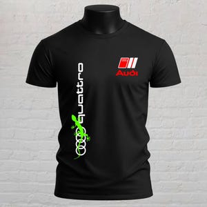 May include: A black short-sleeved t-shirt featuring the Audi logo and the word quattro in white text. A bright green gecko graphic is positioned vertically over the white Audi rings logo on the left side of the chest area.