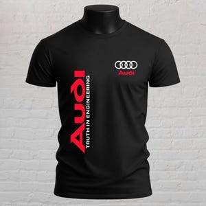 May include: A black short-sleeved t-shirt featuring the white Audi four-ring logo and red Audi text on the upper chest. A large vertical red Audi logo and the white text Truth in Engineering are printed along the left side of the torso.