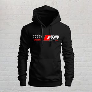 May include: A black long-sleeve hooded sweatshirt featuring the Audi logo in silver and red, alongside the Audi Sport R8 logo in white, red, and black. The garment includes a front kangaroo pocket, a drawstring hood, and ribbed cuffs and hem.