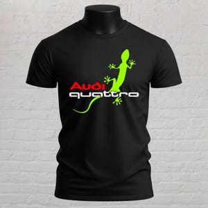 May include: A black short-sleeved t-shirt featuring a bright lime green gecko graphic. The design includes the red text Audi and the white text quattro positioned horizontally across the chest area, centred on the front of the garment.