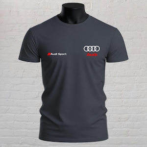 May include: A dark grey short-sleeved t-shirt featuring the white Audi four-ring logo centred on the chest. To the left, the text Audi Sport appears in white next to a small red rectangle, and the red Audi logo is positioned below the rings.