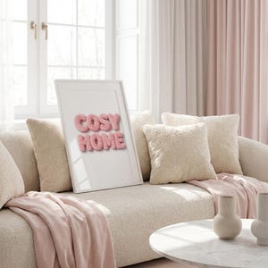 Op de afbeelding: Een gezellige woonkamer met een crèmekleurige bank, zachte kussens en een roze deken. Een ingelijste print met de tekst "COSY HOME" in roze letters leunt tegen de kussens. Roze gordijnen en een wit raam zijn zichtbaar op de achtergrond.
