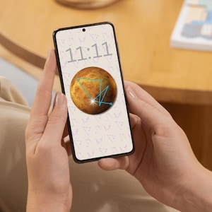 May include: A smartphone displaying a screen with the time 11:11 in gray numerals above a brown planet graphic with blue geometric lines. The phone is held in two hands, with a blurred background.