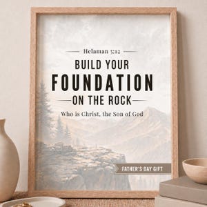 Puede incluir: Impresión artística enmarcada con un marco de madera clara. La impresión presenta el texto "BUILD YOUR FOUNDATION ON THE ROCK" sobre una imagen de paisaje de montañas y árboles. El texto "Helaman 5:12" está en la parte superior, y "Who is Christ, the Son of God" está en la parte inferior. Las palabras "FATHER'S DAY GIFT" también están incluidas.