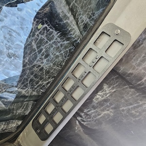 May include: A dark gray metal panel with rectangular cutouts and the text "TWIN PEAKS 4X4 LLC". The panel is attached to a light gray surface with a single screw. The background includes a dark, textured material.