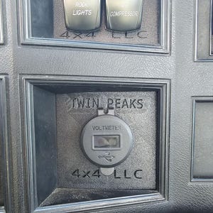 May include: Close-up of a black dashboard panel with the text "TWIN PEAKS" above a round voltmeter with a USB port symbol. The panel also features rocker switches labeled "ROCK LIGHTS" and "AIR COMPRESSOR", and the text "4x4 LLC".