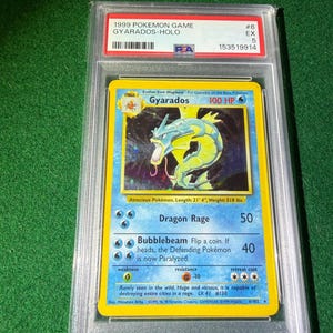 May include: A graded 1999 Pok&eacute;mon card featuring Gyarados-Holo. The card has a blue border with a holographic image of Gyarados. The card is encased in a protective plastic case with a grading label.