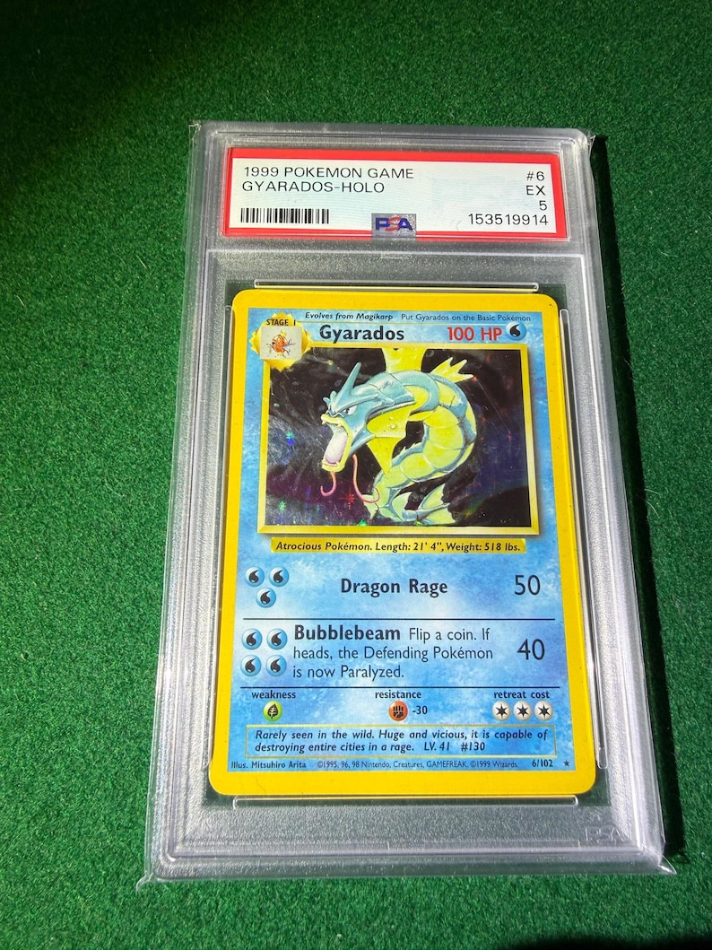 May include: A graded 1999 Pok&eacute;mon card featuring Gyarados-Holo. The card has a blue border with a holographic image of Gyarados. The card is encased in a protective plastic case with a grading label.