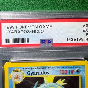May include: A graded Pok&eacute;mon card featuring Gyarados, a Stage 1 Pok&eacute;mon. The card is encased in a clear plastic holder with a red label that reads "1999 Pokemon Game Gyarados-Holo". The card is graded EX 5.
