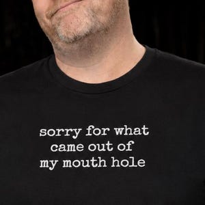 Op de afbeelding: Een zwart t-shirt met korte mouwen en witte tekst op de borst met de tekst: sorry for what came out of my mouth hole. Het shirt heeft een ronde hals en een standaard pasvorm, gemaakt van een effen zwarte stof met witte letters.