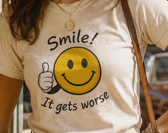 May include: A cream-colored short-sleeve t-shirt featuring a yellow smiley face graphic with a thumbs-up gesture and the black text "Smile! It gets worse" printed on the front, paired with blue denim jeans and a brown leather belt with a round buckle.