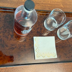 May include: A rectangular serving tray with a brown burl wood pattern, black edges, and two black cylindrical handles. On the tray sits a glass bottle filled with amber liquid, two clear glass tumblers, and a white square napkin with gold trim.