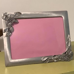 May include: A rectangular silver-toned metal picture frame featuring raised floral and butterfly embellishments at the top-left and bottom-right corners. The frame surrounds a solid pink rectangular insert, providing a clear view of the metallic finish.