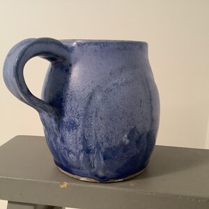 May include: A handmade, blue ceramic mug with a rounded body and a large, curved handle. The mug has a textured surface with darker blue hues at the base, creating a gradient effect. The mug is sitting on a gray surface.