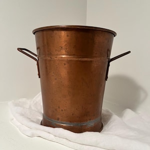 May include: A cylindrical copper bucket featuring a wide rim, a slightly tapered body, and two metal side handles. The exterior surface displays a weathered, metallic finish with visible patina and subtle color variations across the metal.