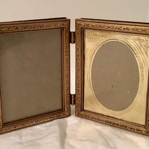 May include: A double-sided folding picture frame made of gold-toned metal. The left side features a rectangular opening, while the right side contains an oval-shaped opening. Both frames are decorated with intricate, embossed scrollwork patterns.