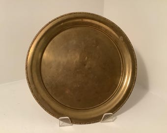 May include: A round, antique brass tray with a decorative edge. The tray has a smooth, reflective surface and a warm, golden hue. The tray is displayed on a clear stand.