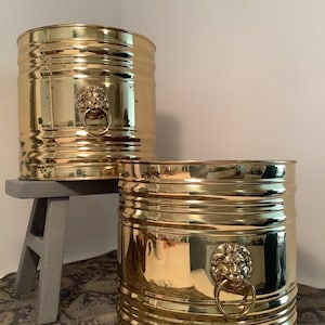 May include: Two gold-colored metal planters with a ribbed design. Each planter features a lion's head with a ring handle. One planter sits on a small gray wooden stool, while the other rests on a patterned surface.
