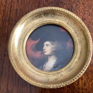 May include: A small, round portrait of a person in a dark hat and blue clothing, set within a gold-colored, ornate frame. The portrait has a red background and is set against a dark wood surface.