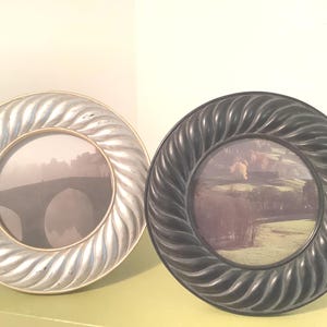 May include: Two circular picture frames with a twisted, rope-like border design. One frame is finished in a metallic silver tone, while the other is finished in a matte black tone. Each frame contains a circular photograph of a landscape scene.