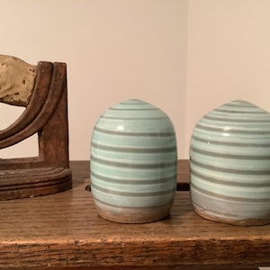 May include: Two dome-shaped ceramic shakers featuring horizontal light blue and grey stripes. The shakers have a smooth, glossy finish on the upper sections and a natural, unglazed clay base. They are positioned side-by-side on a wooden surface.
