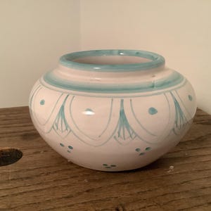 May include: A round ceramic bowl with a white base and a light blue hand-painted pattern. The design features a series of arched lines and small dots encircling the body, with a solid light blue band around the rim and the upper neck area.