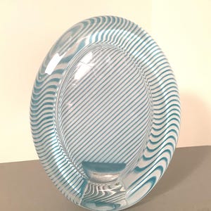 May include: An oval-shaped glass sculpture featuring a clear center with diagonal white stripes and a thick, rounded border decorated with wavy, alternating teal and white lines that create a distinct visual pattern throughout the object.