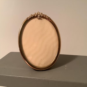 May include: An oval-shaped picture frame with a metallic gold finish. The frame features a decorative, ornate floral-inspired crest at the top center. The interior contains a light cream-colored insert with a subtle, wavy, vertical pattern.