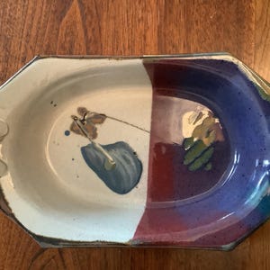 May include: Handmade ceramic serving dish with a unique octagonal shape. The dish is divided into two sections, one cream-colored and the other with a red and blue color scheme. It features abstract floral designs and handles on each side.