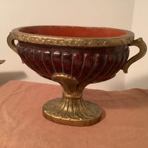 May include: An oval-shaped decorative urn featuring a deep burgundy, mottled exterior with vertical fluting. The rim, two side handles, and the pedestal base are finished in a metallic gold with ornate, embossed detailing. The interior is a solid orange.