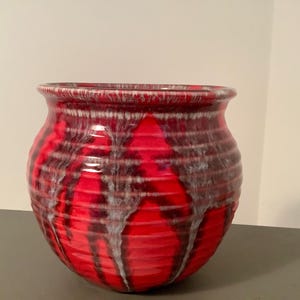 May include: A round, ceramic vase with a wide opening. The vase is primarily red with dark brown and white dripped patterns. The rim is a darker shade of red. The vase is sitting on a gray surface.