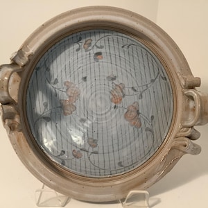 May include: A round, ceramic serving dish with a light blue interior featuring vertical stripes and floral designs in orange and gray. The dish has a beige rim and handles, and is supported by a clear stand.