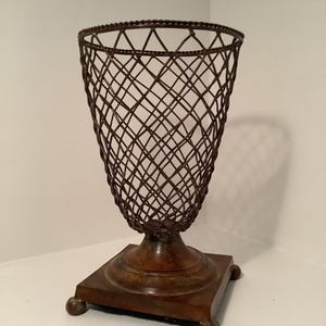 May include: A decorative bronze-colored metal basket with a lattice design. The basket sits atop a square base with rounded feet. The basket's openwork design allows for visibility of the contents.