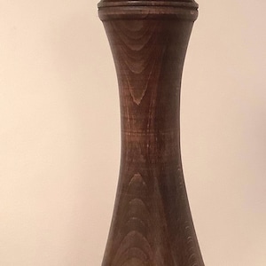 May include: A tall, dark brown wooden pepper mill with a smooth, hourglass-shaped body and a visible wood grain texture. The top features a small, circular silver-colored metal adjustment knob centered on the rounded cap of the grinder.