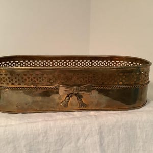 May include: An oval brass planter featuring a decorative lattice pattern along the upper rim, a twisted rope border detail, and a central bow motif with two small hanging tassels on the front side of the polished metallic container.