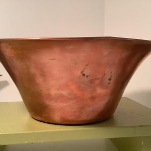 May include: A large, wide-mouthed bowl crafted from polished copper with a distinct hammered texture across its exterior surface. The metal displays a warm, reddish-brown metallic sheen with subtle variations in tone and small dark spots.