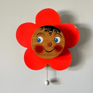 May include: A wall-mounted wooden decoration shaped like a bright red five-petal flower. In the center is a circular wooden face with blue eyes, black curly hair, a small nose, and a smiling mouth. A thin string with a small white bead hangs below.