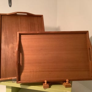May include: Two rectangular wooden serving trays with a warm brown finish and visible wood grain. Each tray features integrated cutout handles on the shorter sides and a raised border around the perimeter for stability and containment.