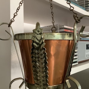 May include: A hanging planter featuring a polished copper bucket body held by a brass-toned metal frame. The frame includes three vertical supports with a leaf-like texture and circular base accents, suspended by three thin metal chains.