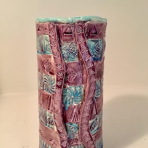 May include: A tall, cylindrical ceramic vase with a textured surface. The vase features a patchwork design in shades of purple and teal, with embossed Celtic knot and geometric patterns. The top rim is a light blue color.
