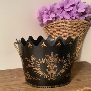 May include: A black metal bucket with a scalloped top edge and two gold-toned side handles. The exterior features a decorative gold-colored floral pattern painted on the front. The bucket has a circular base and a smooth, dark finish.