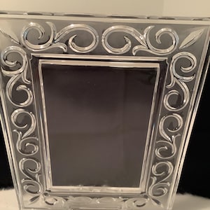 May include: A rectangular clear crystal picture frame featuring an intricate etched scrollwork pattern along the border. The frame includes a small blue and white label at the bottom center with the text Marquis Waterford Crystal printed on it.