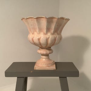 May include: A peach-colored, ornate urn with a scalloped rim, set on a pedestal base. The urn is displayed on a small, gray table. The design features a classic, vintage aesthetic, suitable for home decor.