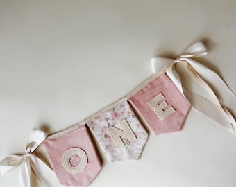 May include: A fabric banner featuring three pennants spelling the word ONE. The first and third pennants are solid dusty pink with cream-colored letters, while the center pennant has a floral pattern with a cream letter. White satin ribbons tie the ends.