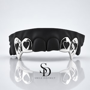 May include: A set of black dental grills featuring silver-toned accents on the canine and lateral incisor teeth. The silver accents display heart-shaped cutouts and a textured diamond-cut pattern. The text SD SMILE DISTRICT is visible below.