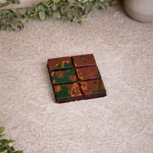 May include: A six-piece wax melt with a marbled effect, predominantly brown with green and gold detailing. The wax melt is rectangular and sits on a neutral, textured surface. Greenery is visible in the background.
